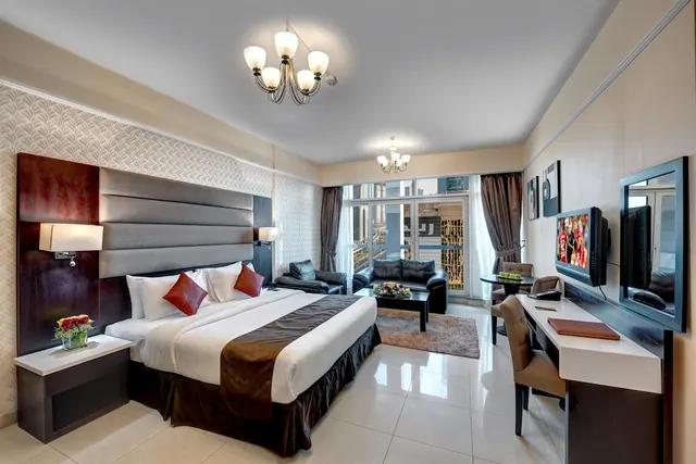A hotel room with a king bed, a sofa and armchairs by glass balcony doors looking onto buildings, a wall-mounted TV and a desk with chairs.