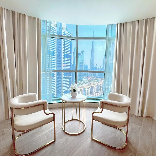 Two cream armchairs with gold frames and a small round side table with a white orchid sit in front of a curved floor-to-ceiling window showing the Burj Khalifa and surrounding towers.