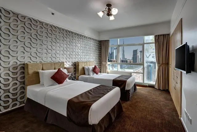 A bedroom with two single beds with red cushions, a patterned feature wall, a wall-mounted television and a window overlooking city towers including the Burj Khalifa.