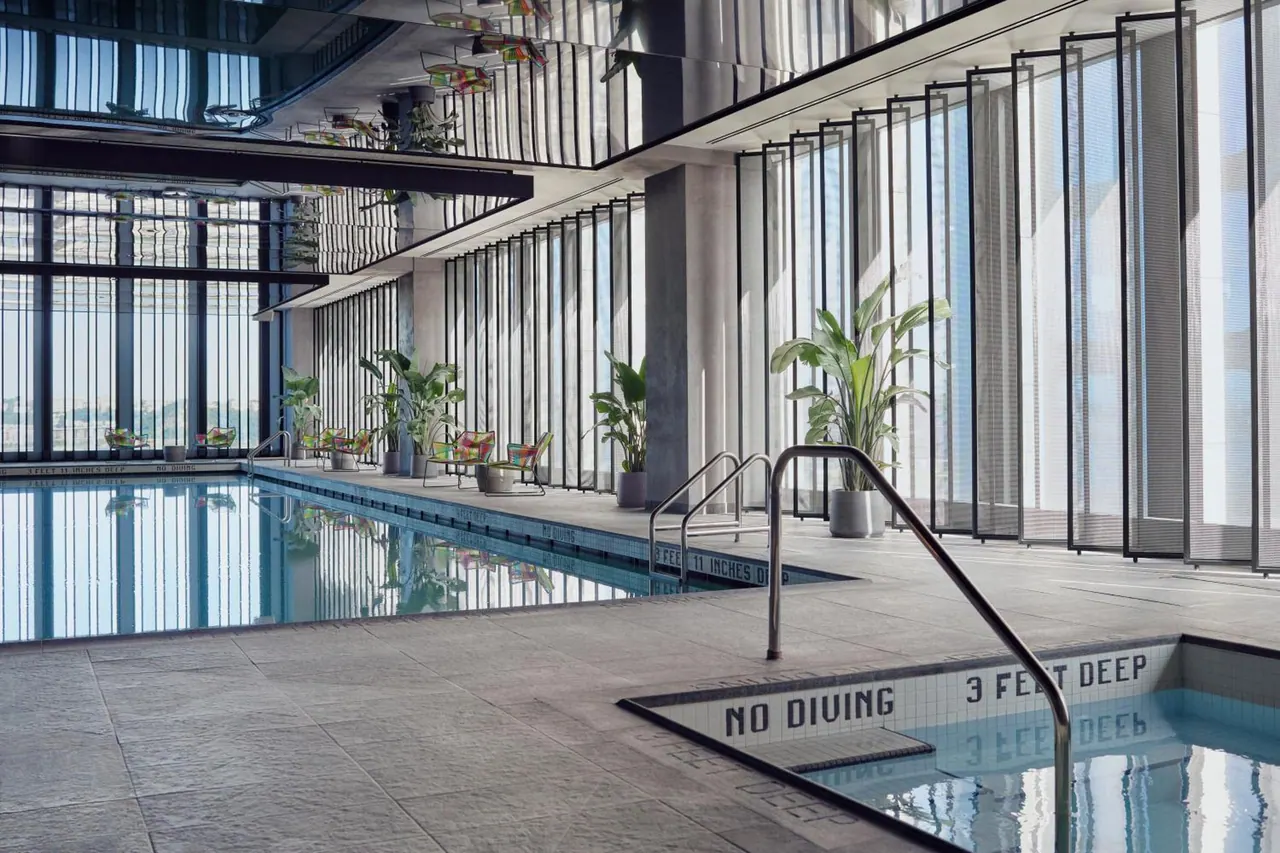 An indoor pool with a tiled edge bearing no-diving and depth signs, with potted plants and lounge chairs beside floor-to-ceiling windows showing hazy city buildings.