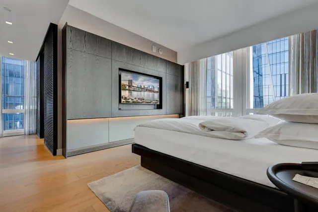A modern hotel bedroom with a king bed, a wall-mounted television and floor-to-ceiling windows looking onto neighbouring glass buildings.