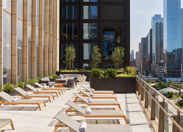 Rooftop terrace with rows of sun loungers with folded towels, planters and lounge seating, with city skyscrapers visible beyond the glass railing.