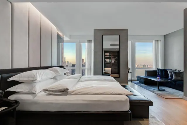 A bedroom with a large made bed, a dark sofa seating area, and floor-to-ceiling windows showing a city skyline with the Empire State Building in the distance.