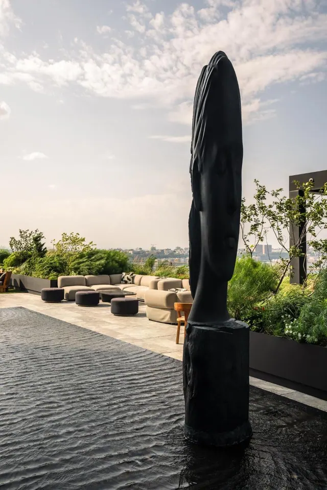 A tall black sculpture of a face stands beside a dark rippled water feature on a rooftop terrace with beige sofas, planters and a hazy city skyline behind.