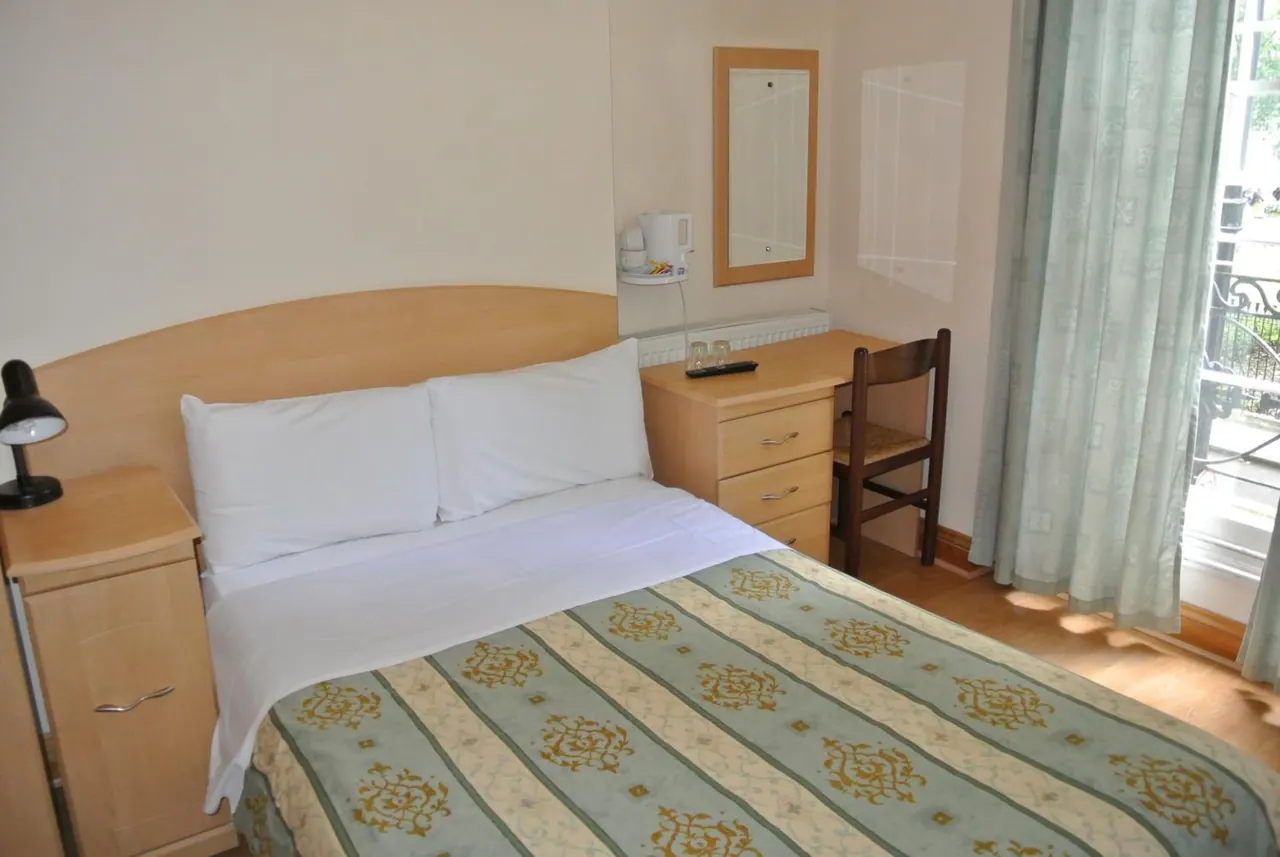 A double bed with white pillows and a patterned green cover sits beside a bedside cabinet, with a dressing table, kettle and chair opposite and a window with curtains at right.