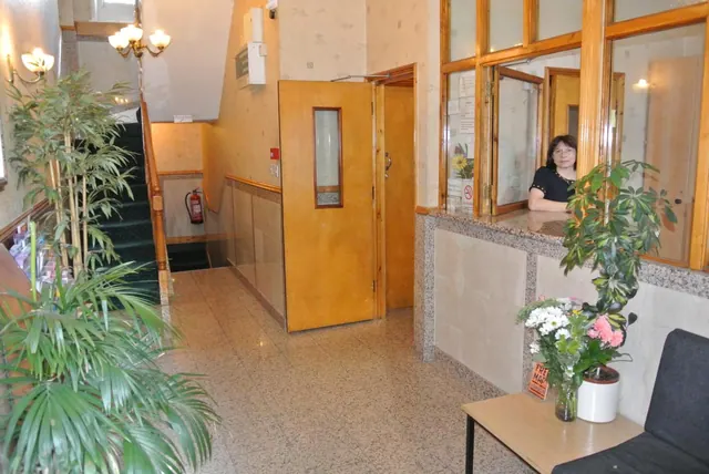 A hotel lobby with a glass reception window where a woman sits behind the counter, with a staircase, potted plants, a small table with flowers and a grey sofa visible.