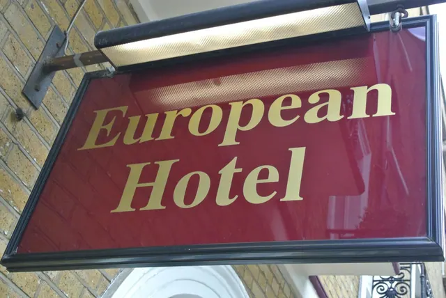 A hanging red sign with gold lettering reading European Hotel is mounted on a brick wall beneath a light fixture.