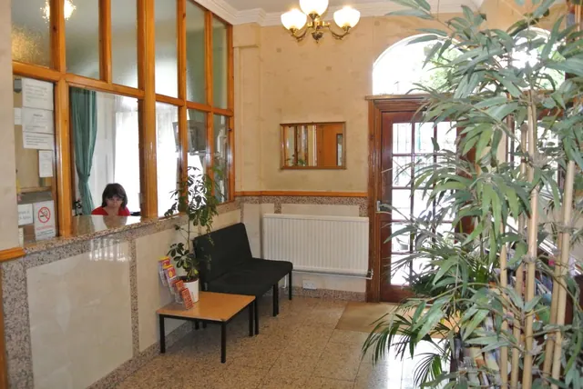 A reception area with a glass-fronted desk where a receptionist sits, a black bench seat, a wooden side table with leaflets, a radiator and a tall potted plant.