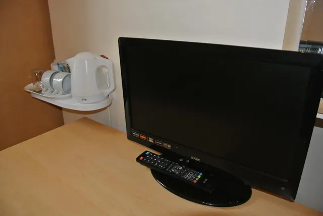 A black flat-screen television with a remote control sits on a wooden surface beside a wall shelf holding a white kettle and cups.