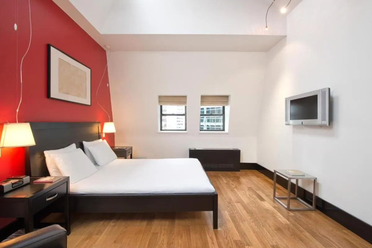 A bedroom with a red accent wall, a king bed with white linens, a wall-mounted television and a wood floor.
