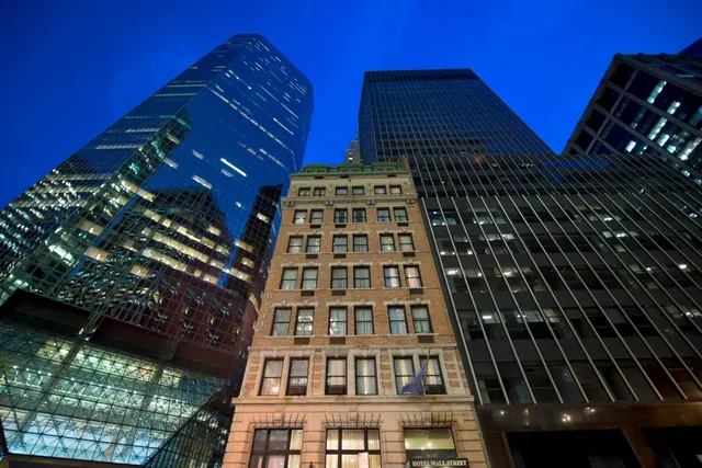 Low-angle view of a brick hotel building with a Hotel Wall Street sign at its base, flanked by tall glass office towers under a deep blue dusk sky.