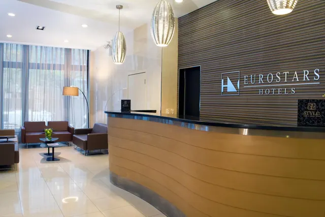 A hotel lobby with a curved wooden reception desk bearing the Eurostars Hotels sign, a seating area with brown armchairs and a coffee table, and pendant lights overhead.