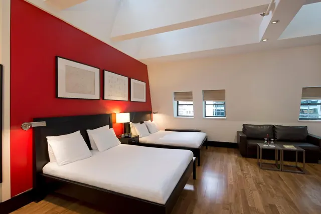 A bedroom with two double beds, a red accent wall, framed prints, a leather sofa and a wooden floor.