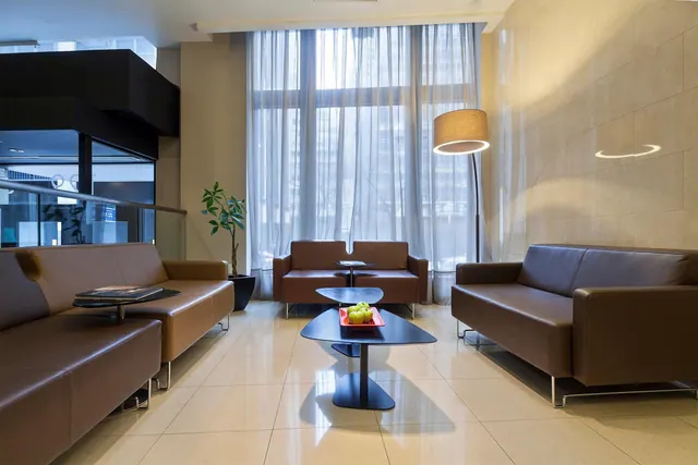 A lounge area with brown leather sofas, a dark coffee table holding a bowl of fruit, a floor lamp, and sheer curtains over tall windows.