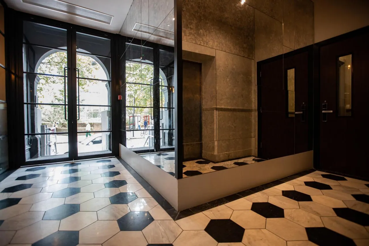A lobby with a black and white hexagonal marble floor, glass doors opening onto a street with trees, and a lift set into a stone-panelled wall.