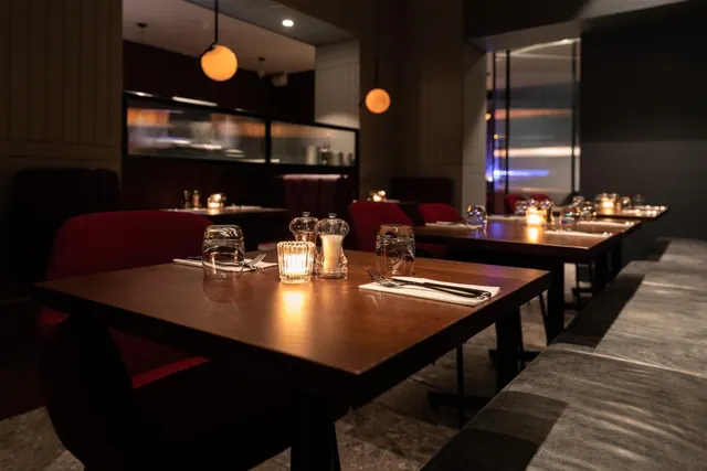 An empty restaurant dining room with set tables, lit candles, red upholstered chairs and a grey banquette along the right wall.