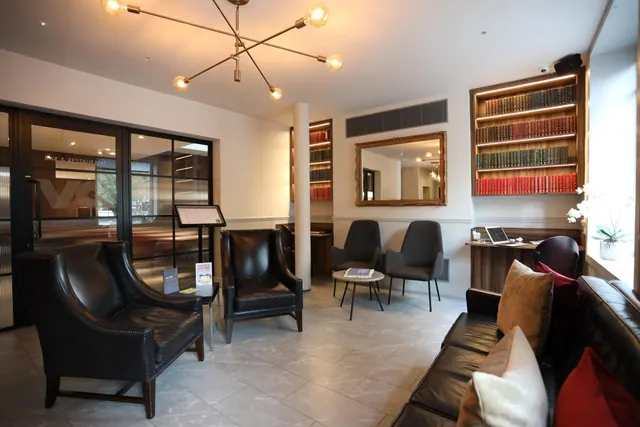 A lounge area in a hotel lobby with leather armchairs, a leather sofa, grey chairs, a framed mirror, bookshelves and a pendant light.