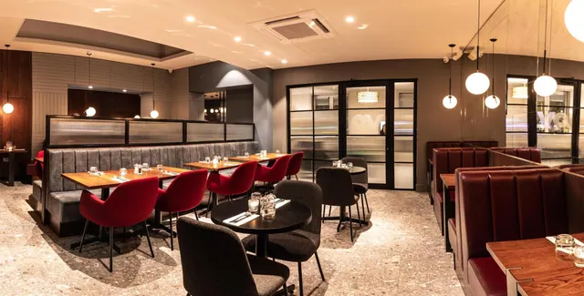 A restaurant dining room with red upholstered chairs, a grey banquette, red leather booths and pendant lights, with no people visible.