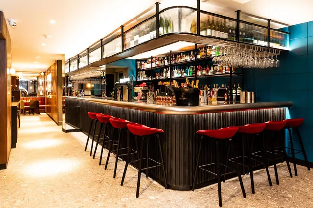 A curved bar counter with red stools, shelves of bottles and hanging wine glasses, and a red banquette seating area in the background.