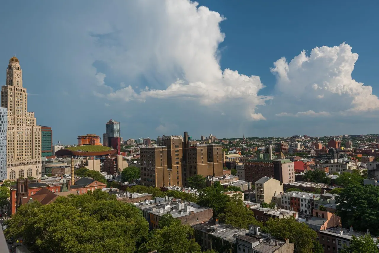 Elevated view over a dense city neighbourhood with a tall gold-topped clock tower on the left under large cumulus clouds.