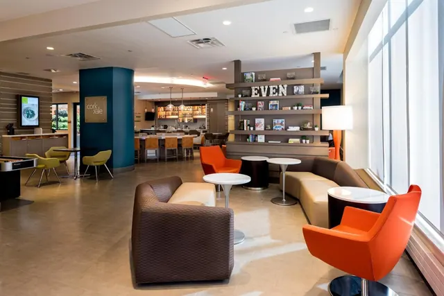 A lobby lounge with brown and cream sofas, orange swivel chairs, round white tables and a bookshelf reading EVEN, with a bar visible in the background.
