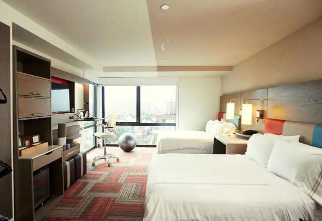 A hotel room with two beds, a wall-mounted television, a desk with a chair, and a window overlooking city buildings.