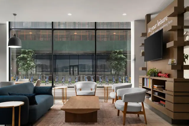 A lounge seating area with a blue sofa, grey armchairs and a wooden coffee table sits in front of large windows overlooking a street with bike docks and trees.