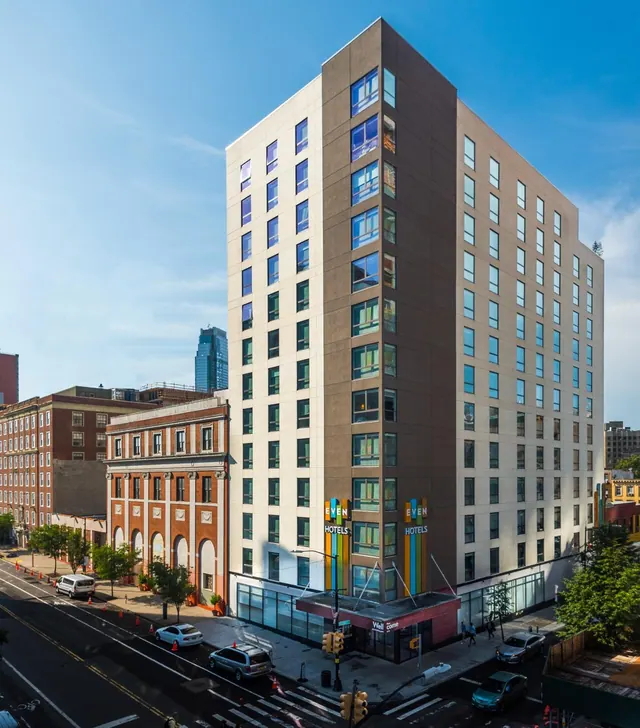 Exterior of a tall hotel building with EVEN Hotels signage at street level, beside a brick building, parked cars and a traffic-lit intersection under a clear blue sky.