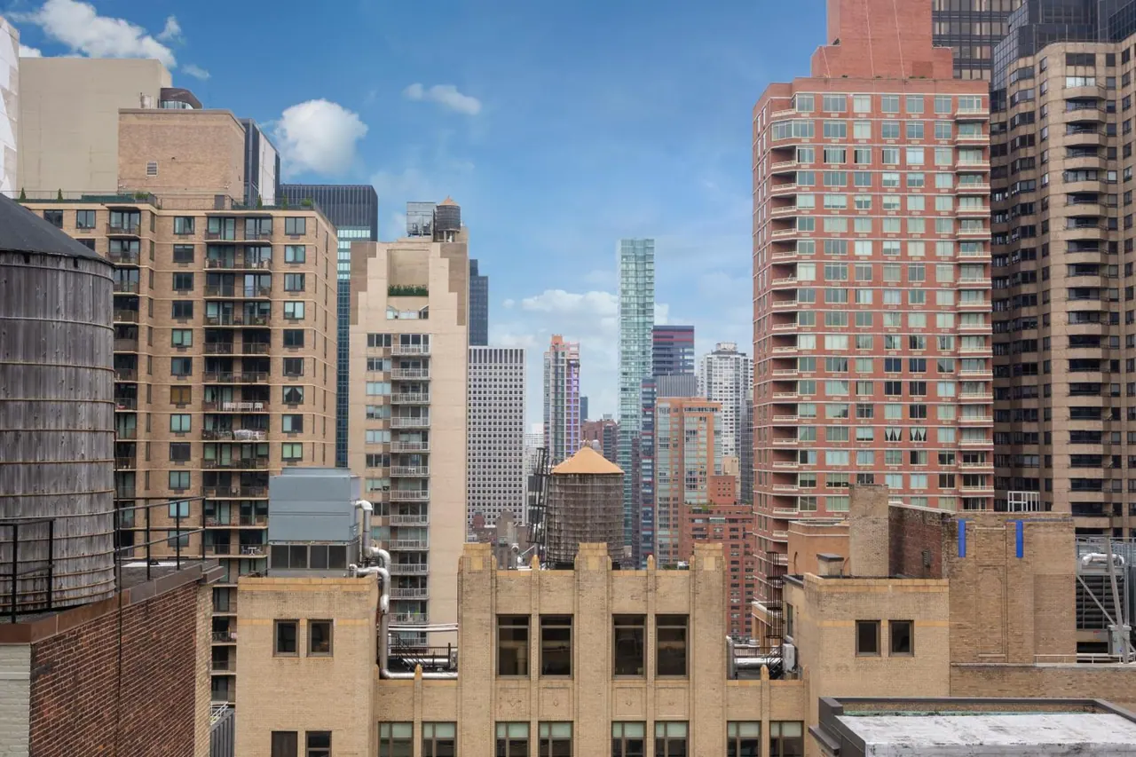 A view over rooftops and high-rise towers under a partly cloudy blue sky, with a wooden water tower visible on the left.