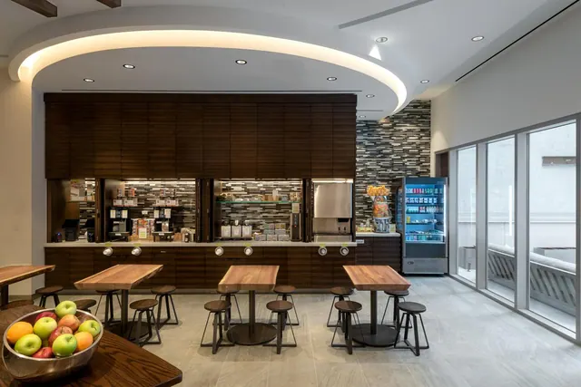 A dining area with a coffee and snack station, wooden tables and stools, a fruit bowl in the foreground, a drinks fridge, and floor-to-ceiling windows on the right.