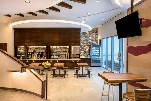 A hotel breakfast area with a serving counter, coffee machines, wooden tables and stools, a wall-mounted television, and a bowl of apples on a table.