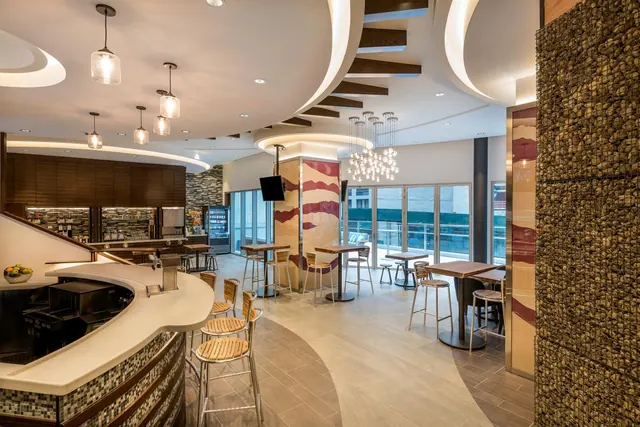 A modern hotel lounge with a curved bar counter, pendant lights, wooden bar stools and high tables beside floor-to-ceiling windows.