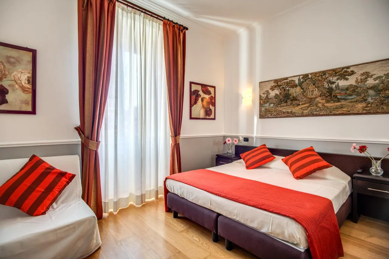 A bedroom with a double bed under a red throw and red striped cushions, a white sofa on the left, red curtains by a window, and a tapestry on the wall.