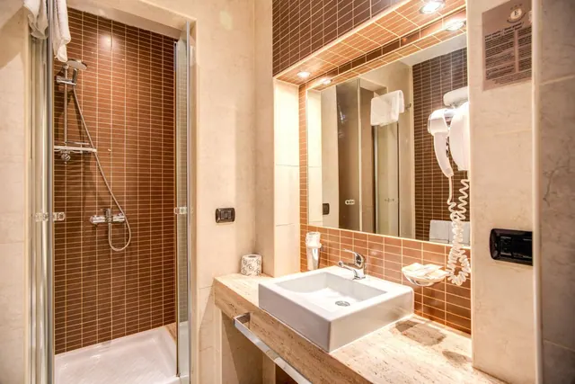 A bathroom with a glass-enclosed walk-in shower, brown tiled walls, a square basin on a stone countertop, and a mirror with a wall-mounted hairdryer.