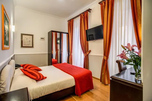 A double bed with red bedding and striped pillows faces a wall-mounted television, a mirrored wardrobe and a window with orange curtains, with a vase of pink lilies on a dark wooden desk.