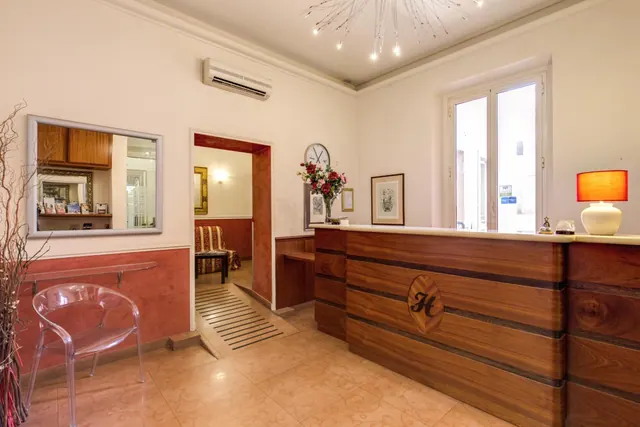 A hotel reception area with a wooden front desk, a red table lamp, a wall-mounted air conditioner and a doorway leading to a seating area with a striped armchair.