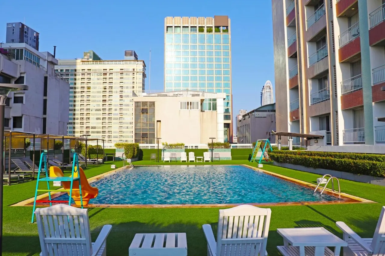 A rectangular swimming pool on an artificial turf deck with white chairs, a children's slide and a view of city high-rises under a clear blue sky.