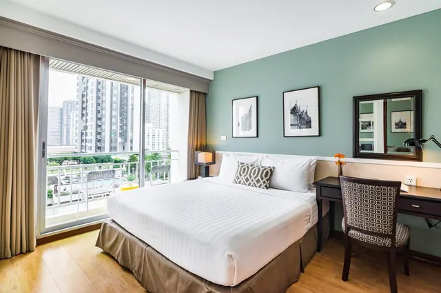A bedroom with a made king bed, a work desk with a chair, and a sliding glass door opening onto a balcony overlooking high-rise buildings.