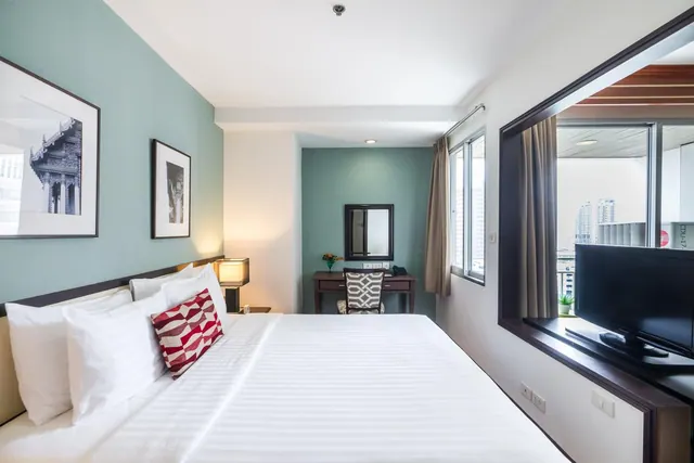 A bedroom with a made king bed, framed prints on a teal wall, a desk and chair, and a wall-mounted television beside a window showing city towers.