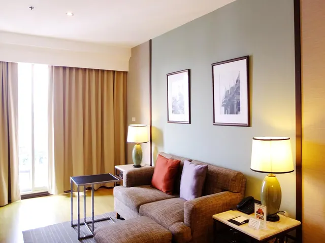 A hotel living area with a grey sofa and two cushions, table lamps on side tables, framed prints on a pale green wall, and curtains beside a window.