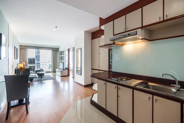An open-plan apartment room with a kitchenette and sink in the foreground, a dining table and sofa beyond, and sliding glass doors leading to a balcony.