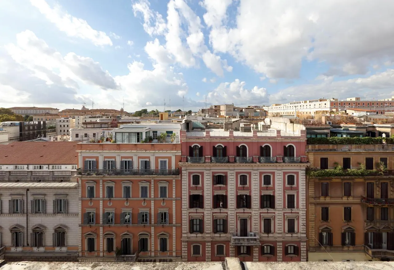 View over rooftops and building facades in a Rome neighbourhood under a partly cloudy sky, with a terrace edge in the foreground.