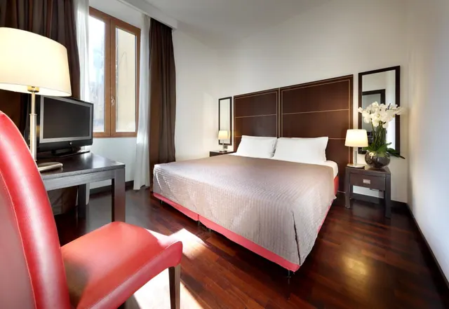 A double bed with a dark wood headboard, bedside lamps and a flat-screen television on a desk, with hardwood floors and a red leather armchair in the foreground.