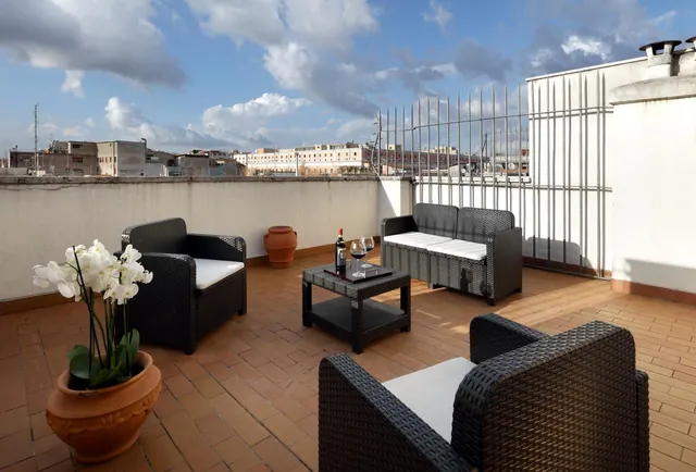 A rooftop terrace with wicker armchairs and a sofa, a coffee table holding a wine bottle and glasses, a potted white orchid, and a view over surrounding city buildings under a partly cloudy sky.