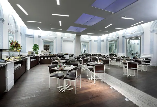 A dining room with rows of tables and dark-brown chairs, a buffet counter along the left wall, and arched windows with mirrors on the right.