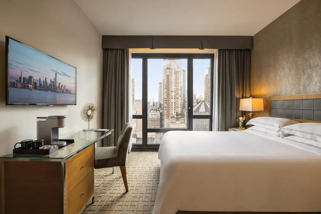 A hotel bedroom with a king bed, a desk with a coffee machine and mugs, a wall-mounted television, and a window overlooking city buildings.