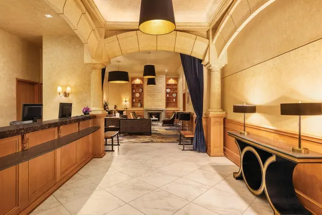A hotel lobby with a wood-and-granite reception desk with two monitors, an arched stone ceiling, pendant lamps, a console table with two table lamps, and a seating area with a fireplace in the background.