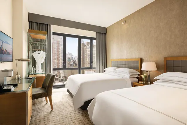 A hotel bedroom with two beds facing a window that looks out onto city high-rises, with a desk, chair and wall-mounted television.