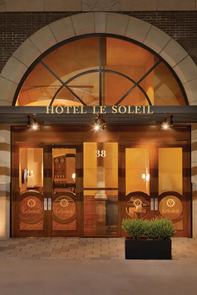 The entrance of Hotel Le Soleil at dusk, with an arched window, an illuminated sign above the doors, and a planter with shrubs on the pavement.