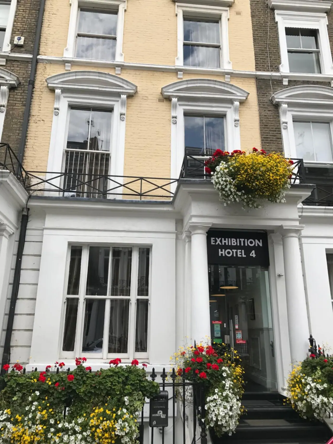 Street-level view of the front of a cream and white terraced building with an Exhibition Hotel 4 sign over the entrance, with flower baskets on the upper balcony and window boxes at the ground floor.